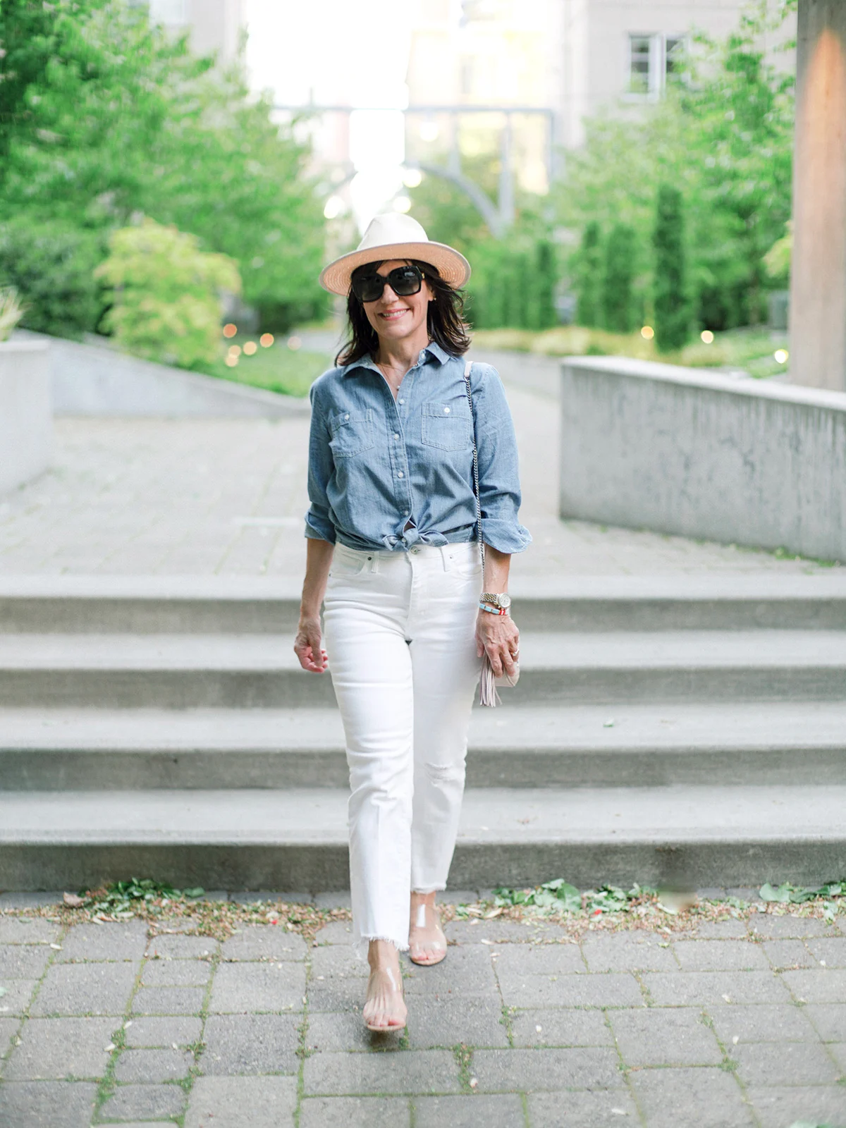 Susie Wright wearing a blue chambray shirt tied at the waist, white cropped jeans, neutral heels and a straw hat.