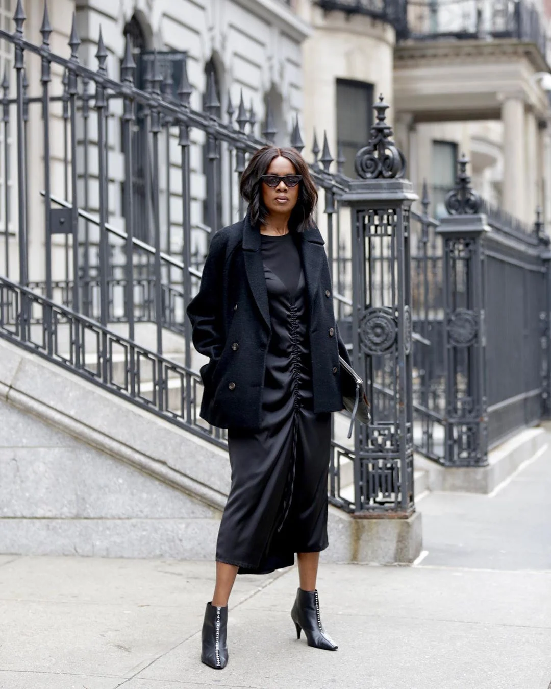 Elaine Davis wearing a black blazer over a gathered black midi dress with black ankle boots.