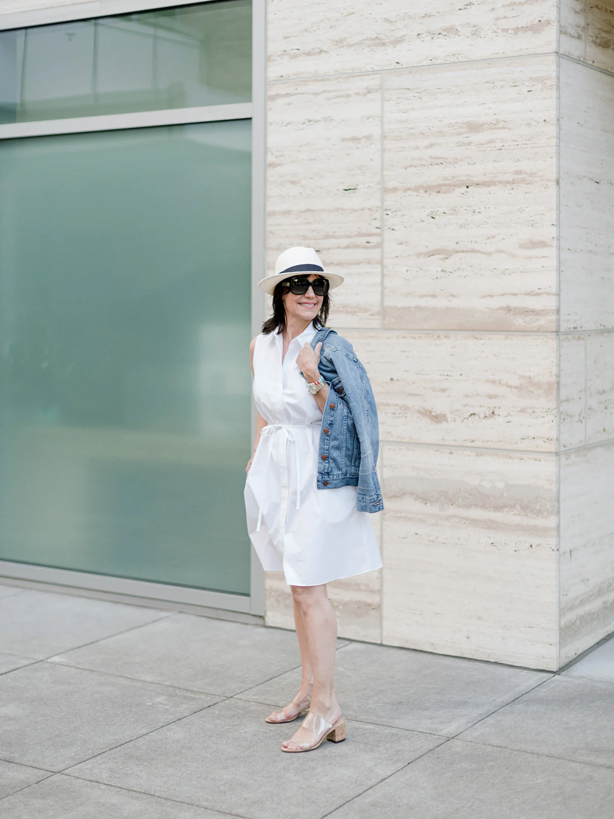 Susie Wright wearing a white shirt dress, a denim jacket over one shoulder, a pale hat and light-colored heels.